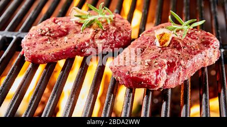 Due crude e spesse bistecche di manzo succose guarnite con grigliate al rosmarino sulle bollenti calette di un fuoco al barbecue in primo piano Foto Stock