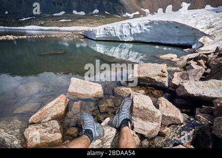 Gambe di uomo in scarpe di tracking e vista del ghiacciaio nevoso sul lago di montagna Foto Stock