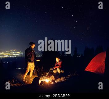 Coppia felice escursionisti caldo hanno le mani in prossimità del fuoco sotto il cielo notturno con le stelle e le luci della città in background. Foto Stock