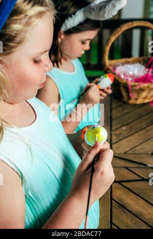 Le ragazze piccole cute stanno dipingendo le uova di Pasqua con i pennelli che rimangono vicino alla tabella di legno nel cortile. Foto Stock