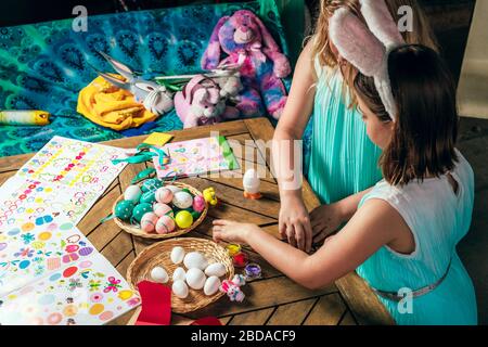 Le ragazze piccole cute stanno dipingendo le uova di Pasqua con i pennelli che rimangono vicino alla tabella di legno nel cortile. Foto Stock