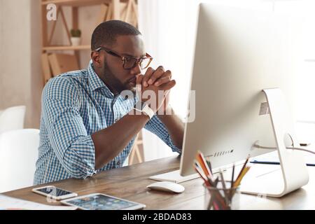 Pensare al business. Giovane afro americano pensieroso in occhiali che guardano lo schermo del computer mentre si siede al suo posto di lavoro a casa. Lavorare in remoto. Freelance. Ambiente di lavoro domestico Foto Stock