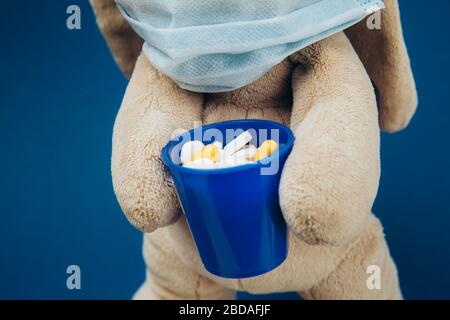 Il bunny giocattolo indossa una maschera medica protettiva. Flapper contiene un bicchiere di pillole. Interrompere il coronavirus. Concetto di medicina. Primo piano. Foto Stock