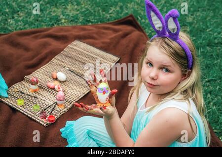 Ragazza carina che indossa le orecchie conigliate il giorno di Pasqua seduta su coperta su erba in giardino e mostrare la sua pittura sull'uovo in mani sporche colorate. Foto Stock