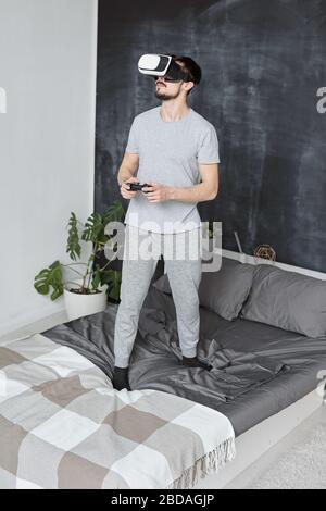 Contenuto bel giovane uomo con la barba in piedi sul letto e utilizzando il joystick durante il gioco video in occhiali di realtà virtuale Foto Stock
