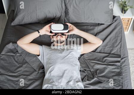 Vista dall'alto di un giovane e rilassato uomo sdraiato a letto e con occhiali di realtà virtuale mentre guardi il film in 3D a casa Foto Stock