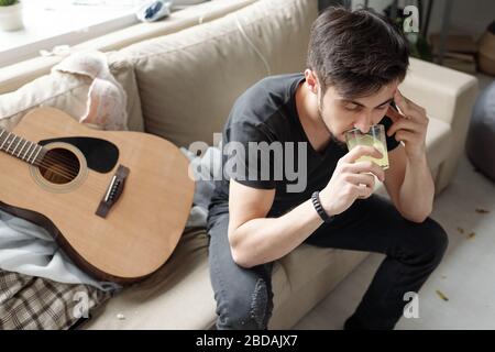 Giovane uomo ubriaco con mal di testa seduto sul divano con chitarra e pillola da bere dissolto in un bicchiere d'acqua dopo la festa Foto Stock