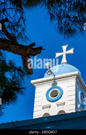 Bella croce bianca della chiesa ortodossa del Monastero di San Nicola, lago Vistonida, Porto Lagos, regione Xanthi, Grecia settentrionale, torre campanaria vista parziale contro il cielo blu chiaro, incorniciato da alberi Foto Stock