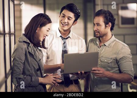 un team di tre giovani imprenditori asiatici che hanno una discussione utilizzando un computer portatile in ufficio felice e sorridente Foto Stock