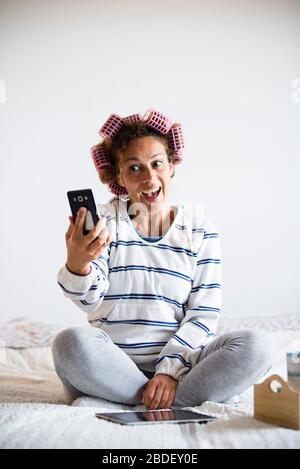 Donna con hairÂ curlersÂ seduta sul letto e prendendo selfie Foto Stock