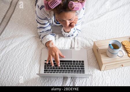 Donna con arricciacapelli sdraiata sul letto e che utilizza un computer portatile Foto Stock