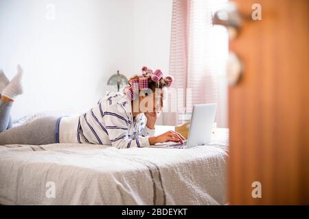 Donna con arricciacapelli sdraiata sul letto e che utilizza un computer portatile Foto Stock