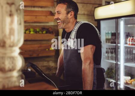 Il lavoratore del caffè è in piedi dietro il banco del check-out e sorridente. Barista con un tovagliolo sulla spalla e grembiule che lavora nel bar. Foto Stock