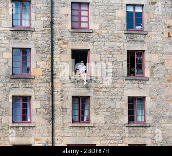 Leith, Edimburgo, Scozia, Regno Unito. 8 Aprile 2020. Covid-19 Lockdown: In uno dei giorni più caldi di sole finora quest'anno con persone in Lockdown. Un uomo e il suo cane siedono precariosamente in una finestra aperta del terzo piano nel magazzino cooperage convertito in appartamenti sulla Shore lungo l'acqua di Leith Foto Stock