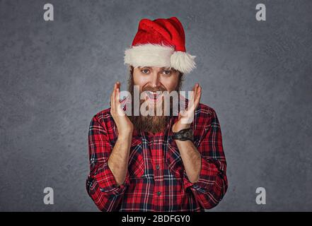 Sorpreso eccitato felice uomo bearded che indossa il cappello rosso di babbo natale che osserva scioccato da che cosa ha visto lo sfondo grigio isolato. Emozione positiva Foto Stock