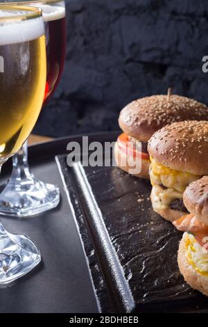 tazza di birra con hamburger e patatine fritte da vicino, bicchieri di birra con birra artigianale scura e leggera con schiuma, su uno sfondo di hamburger, con una varietà Foto Stock