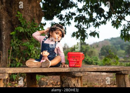 Margao, Goa/India - 2 Aprile 2020 : giocosa ragazza indiana Pretty bambino/neonato indossando un cappuccio, leggendo un libro e giocando avendo campi e tramonto nel backg Foto Stock