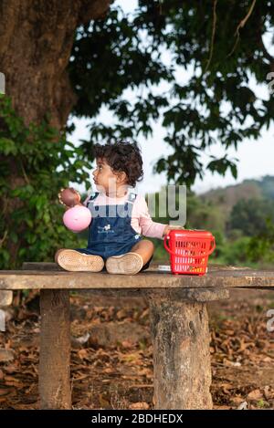 Margao, Goa/India - 2 Aprile 2020 : giocosa ragazza indiana Pretty bambino/neonato indossando un cappuccio, leggendo un libro e giocando avendo campi e tramonto nel backg Foto Stock