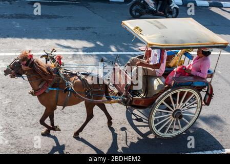 Port Benoa, Bali Indonesia -- Feb 28, 2016. Una carrozza trainata da cavalli e un autista trasportano una donna che indossa un vestito rosa e un cappello a Bali, in Foto Stock