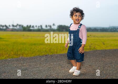 Margao, Goa/India - 2 Aprile 2020 : giocosa ragazza indiana Pretty bambino/neonato indossando un cappuccio, leggendo un libro e giocando avendo campi e tramonto nel backg Foto Stock