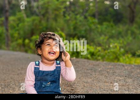 Margao, Goa/India - 2 Aprile 2020 : giocosa ragazza indiana Pretty bambino/neonato indossando un cappuccio, leggendo un libro e giocando avendo campi e tramonto nel backg Foto Stock