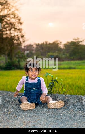 Margao, Goa/India - 2 Aprile 2020 : giocosa ragazza indiana Pretty bambino/neonato indossando un cappuccio, leggendo un libro e giocando avendo campi e tramonto nel backg Foto Stock