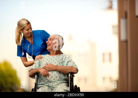 Giovane medico e la sua paziente anziano ridere insieme come lei lo spinge in una sedia a rotelle. Foto Stock