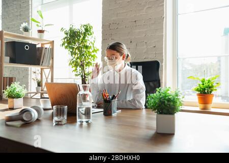 Riunione. Donna che lavora in ufficio da sola durante il coronavirus o la quarantena COVID-19, indossando maschera facciale. Giovane donna d'affari, manager che svolge attività con smartphone, notebook, tablet ha una conferenza online. Foto Stock