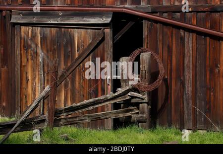 Primo piano di un vecchio, storico stagionato porta fienile di sequoia, laminato filo spinato, e recinzione ringhiera nel giorno di sole. Un po 'di erba verde, diverse linee diagonali. Foto Stock