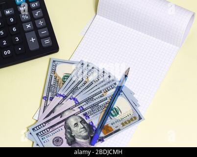 Diverse nuove banconote da cento dollari si trovano su un blocco note vuoto. Calcolatrice e penna su tavolo giallo Foto Stock