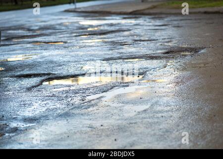 Vicino a una strada in pessime condizioni con grandi buche piene di piscine d'acqua piovana sporche. Foto Stock