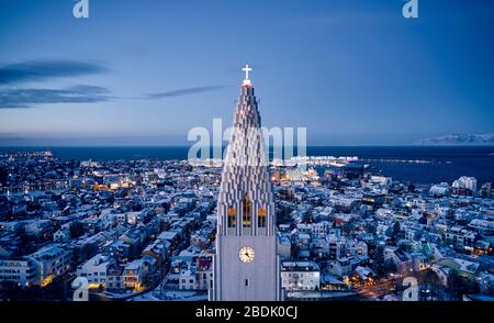 Vista del drone dell'alta torre bianca della chiesa di Hallgrimskirkja con la croce scintillante contro la città illuminata di Reykjavik nel crepuscolo Foto Stock