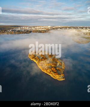 Dal drone sopra vista di piccola e luminosa isola rocciosa coperta di nebbia situata vicino alla costa islandese con città moderna sullo sfondo in un giorno nuvoloso Foto Stock