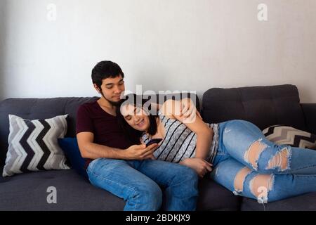 Giovane adulto ispanico sposato coppia abbracciare sul divano guardando uno smartphone sorridente divertirsi a guardare un video Foto Stock