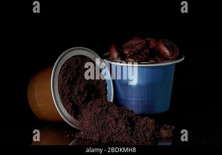 vista orizzontale di due capsule di caffè espresso color oro e blu con chicchi di caffè e versamenti di caffè macinato e loro riflessione su una superficie su b Foto Stock