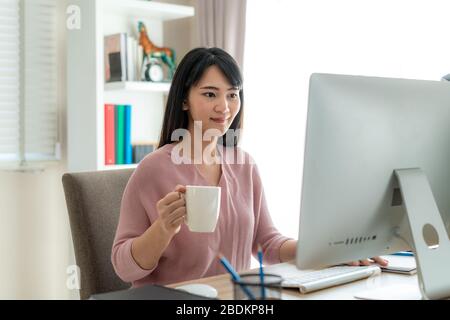 Asian bella giovane donna lavorare da casa lavorare sul computer e bere caffè mentre si lavora in soggiorno a casa. Stile di vita di distanza sociale i Foto Stock