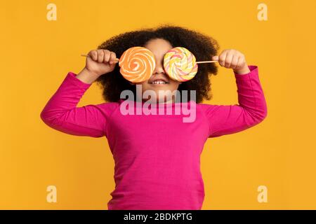 Dente dolce. Divertente bambina nera che copre gli occhi con lollipops colorati Foto Stock