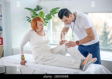 Giovane uomo serio che guarda il ginocchio dei suoi pazienti Foto Stock