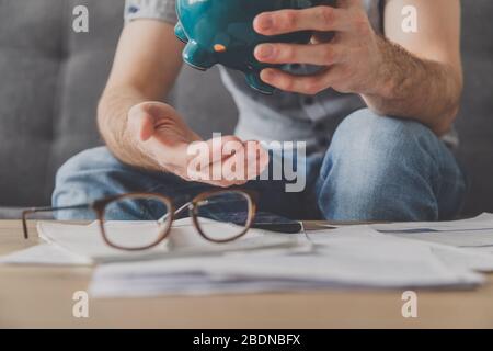 L'uomo seduto ad un tavolo pieno di fatture non pagate scrolla l'ultimo centesimo dalla banca piggy. Spendi i tuoi ultimi risparmi. Disoccupazione, povertà, fallimento Foto Stock