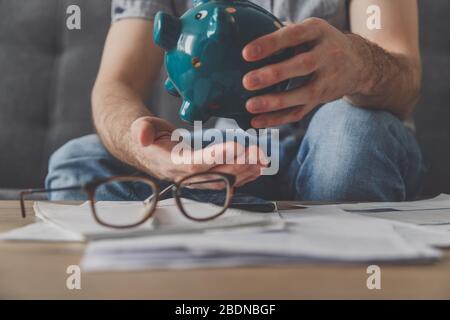 L'uomo seduto ad un tavolo pieno di fatture non pagate scrolla l'ultimo centesimo dalla banca piggy. Spendi i tuoi ultimi risparmi. Disoccupazione, povertà, fallimento Foto Stock