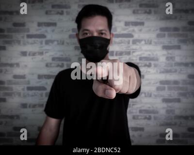 Uomo serio asiatico che indossa maschera facciale protettiva nera e camicia nera che punta il dito alla fotocamera su sfondo scuro, Coronavirus e pm 2.5 Concept. Foto Stock