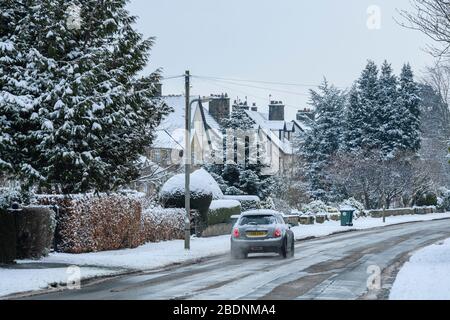 Città invernale innevata (strada coperta di neve, Mini auto che viaggia lungo la tranquilla strada residenziale guidando oltre le case) - Ilkley, Yorkshire, Inghilterra, Regno Unito Foto Stock