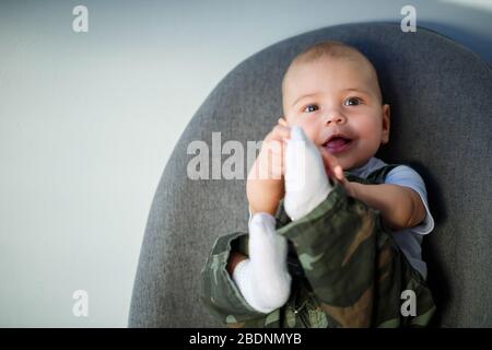 ragazzino di 8 mesi di età in tute, una camicia bianca e calze bianche sedute su una sedia grigia Foto Stock