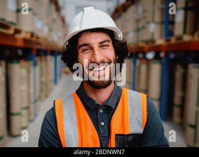 Ritratto di felice lavoratore maschile in magazzino con giubbotto arancione e casco bianco in piedi tra gli scaffali Foto Stock