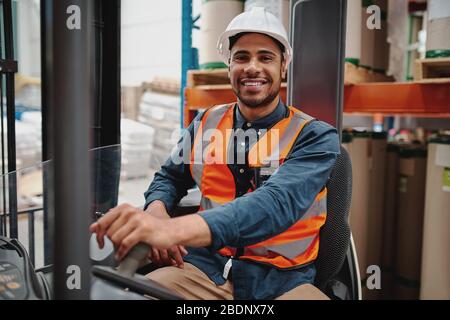 Ritratto di felice conducente che indossa casco bianco e giubbotto seduto in macchina elevatore a forche trasporto merci in magazzino Foto Stock