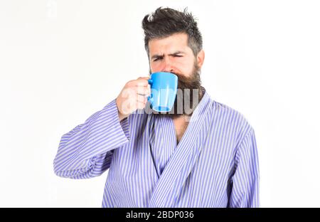 Macho in accappatoio beve tè o caffè con il viso del sonno. Il ragazzo sembra assonnato, ha bisogno di caffè. L'uomo con barba e baffi tiene una tazza di caffè, isolato su bianco. Caffè del mattino Foto Stock