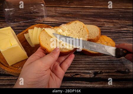 Qualcuno spalma il burro su pane tostato di grano fresco con un coltello, un piatto di burro di legno con un pezzo di burro e fette di formaggio su uno sfondo di legno. Clo Foto Stock