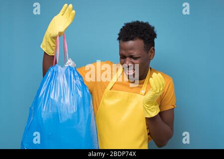 African american maschio in grembiule e guanti in possesso di un sacchetto di spazzatura stinky isolato su sfondo blu Foto Stock