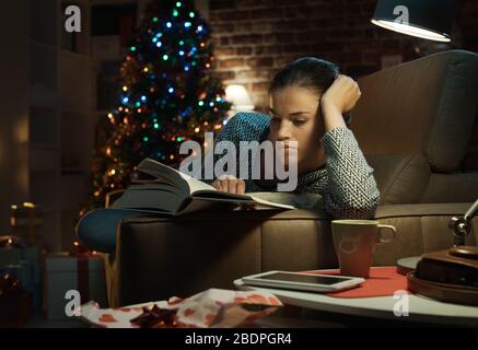 Donna sdraiata sul divano e leggendo un libro che ha ricevuto come regalo di Natale, ha appena sdraiato Foto Stock