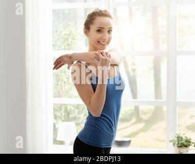 Giovane donna sportiva che si esercita a casa nel soggiorno, si stende il braccio e sorridente alla macchina fotografica Foto Stock